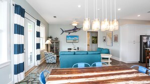 Ein modernes Wohnzimmer mit Esstisch aus Holz, blauen Stühlen, einem türkisgrünen Sofa und einem Deckenventilator.