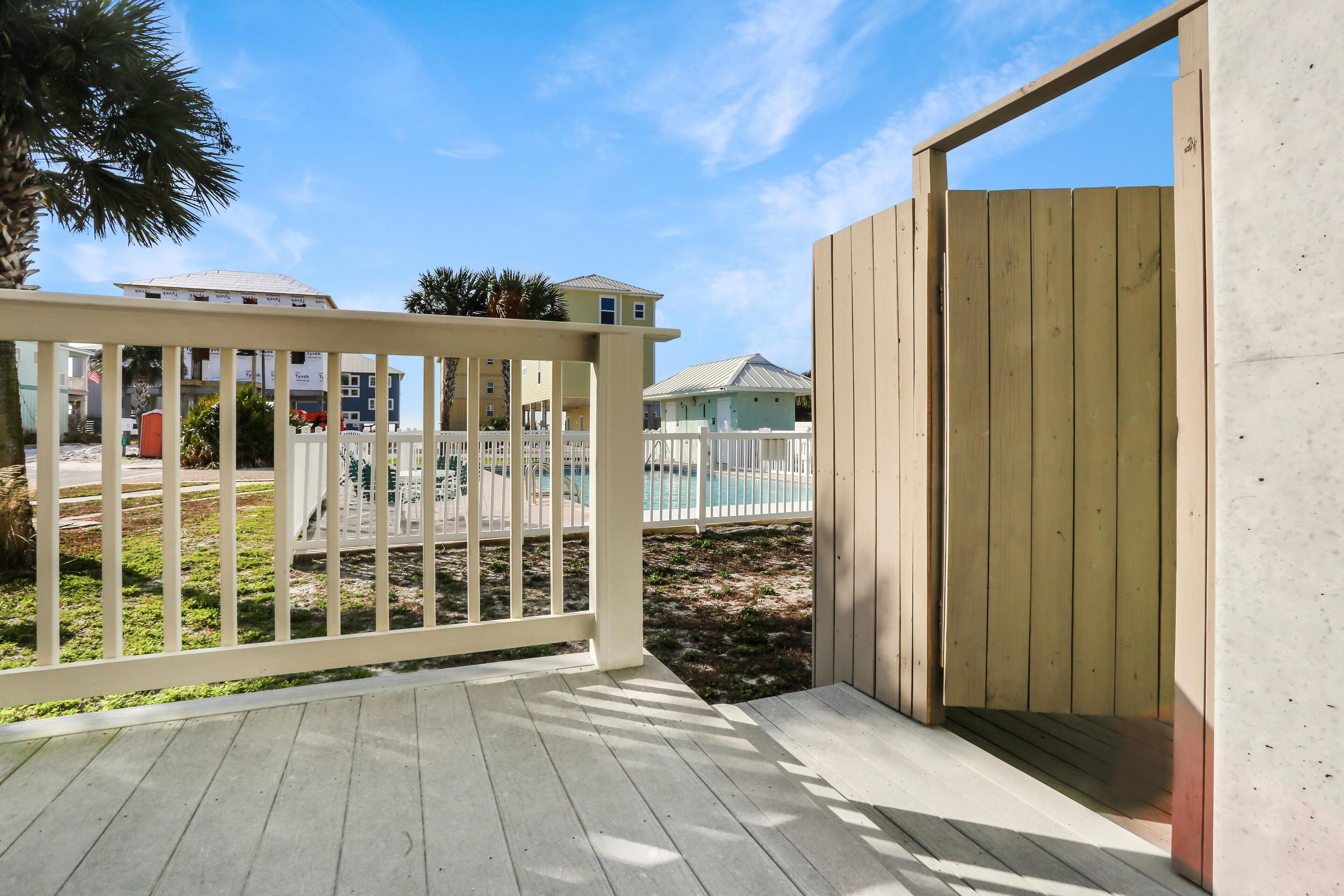 A wooden deck with a white railing, a pool in the background, and a building with a white fence.