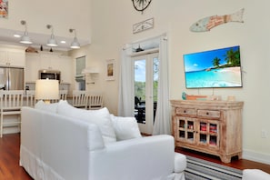 A living room with a white sofa, a wooden cabinet, a television displaying a beach scene, and a door leading to a balcony.