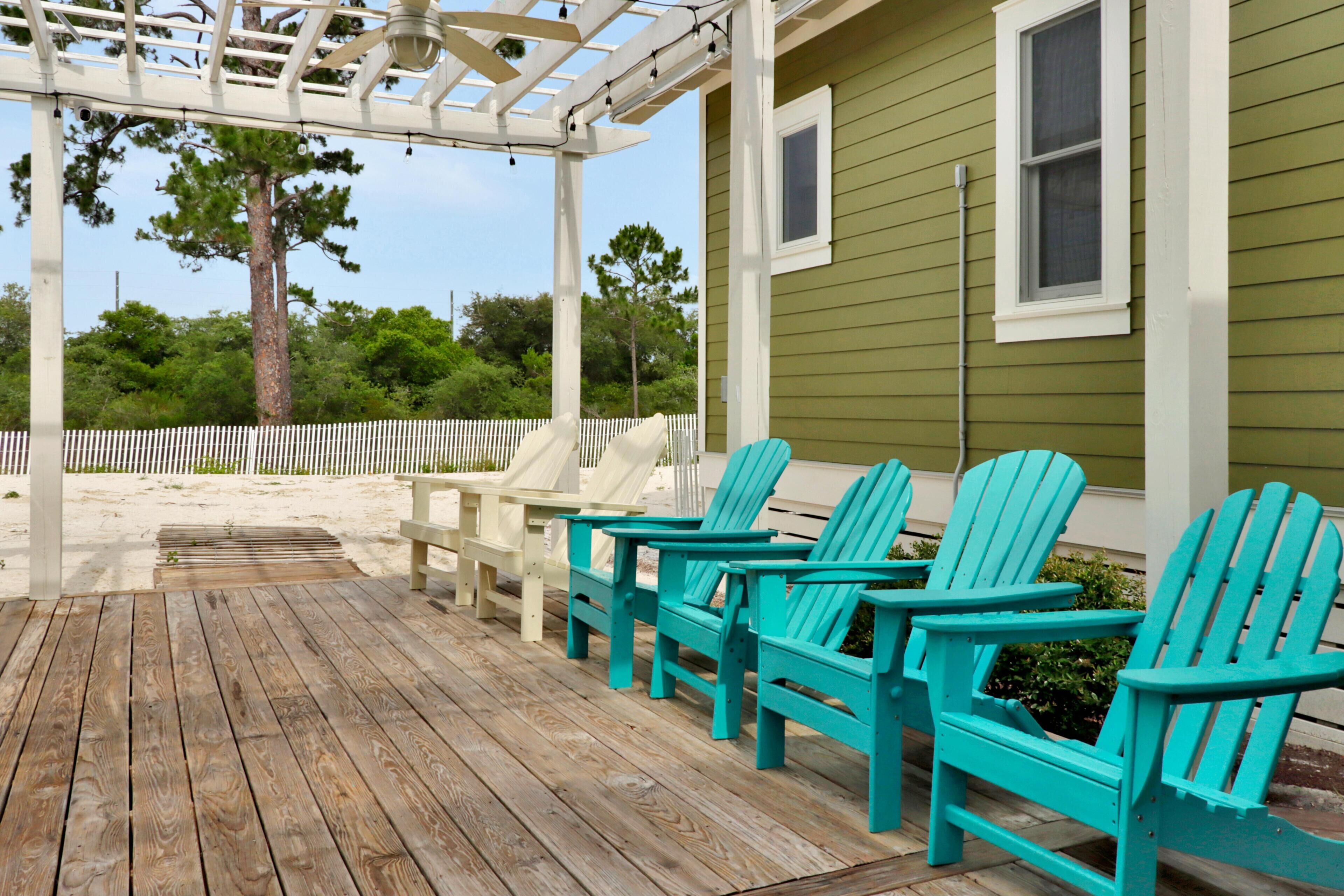 Een houten terras met turquoise Adirondack-stoelen, een witte pergola en een groen gebouw met een wit raam.