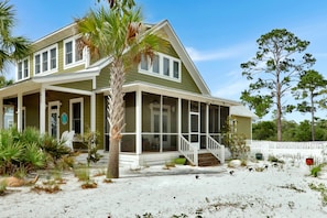 A two-story house with a covered porch, a fenced yard, and a sandy beach in front.