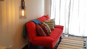 Ein Wohnzimmer mit einem roten Sofa, einem Bild an der Wand, einer Pendelleuchte und einem Fenster mit Vorhängen.