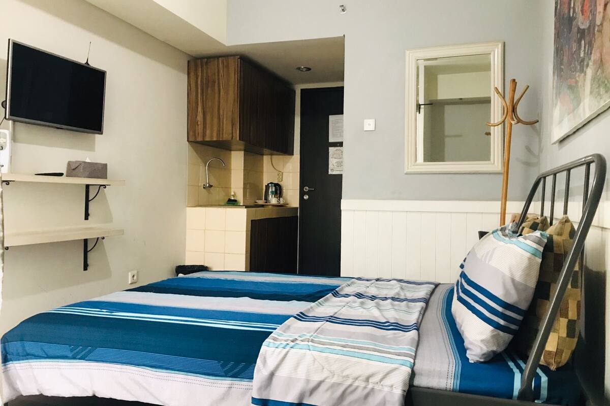 Een slaapkamer met een bed, een aan de muur bevestigde televisie, een keukenhoek en een deur naar een andere kamer.