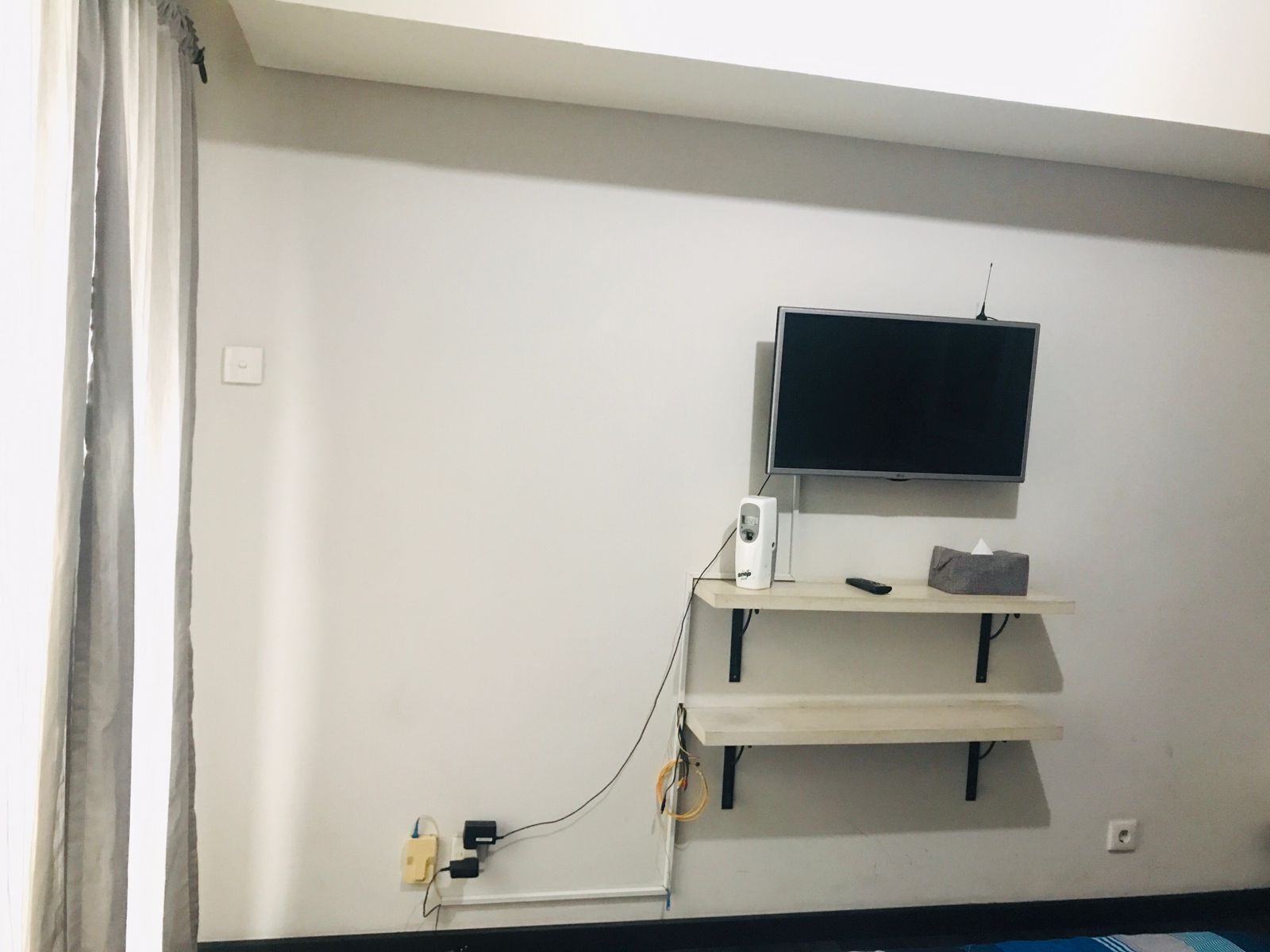 Une pièce avec un téléviseur fixé au mur, une étagère en dessous et une prise électrique avec un câble.