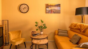 Una sala de estar con un sofá amarillo, una mesa redonda de madera, un sillón amarillo, un reloj de pared, un cuadro y una lámpara de pie.