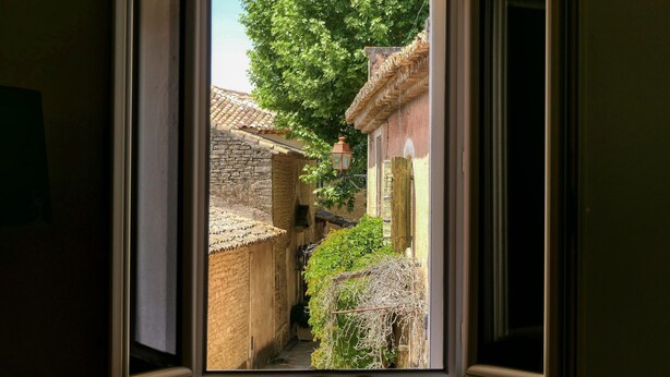 Vista de una calle estrecha a través de una ventana abierta, mostrando arquitectura tradicional con muros de piedra y tejados de tejas.