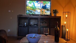 Un televisor sobre un mueble de madera, una botella de vino, una copa de vino y un bol de comida sobre una mesa redonda.