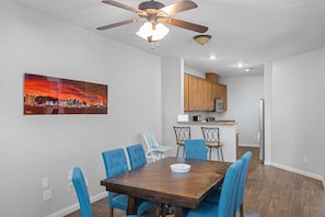 Comedor con mesa de madera y sillas azules, ventilador de techo y un cuadro de paisaje urbano en la pared.
