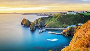 Un paysage côtier caractérisé par des falaises rocheuses, une mer calme et quelques bâtiments situés sur le versant de la colline.