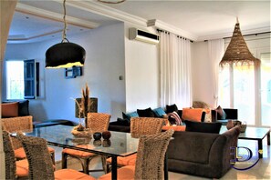 A modern living room with a glass dining table, wicker chairs, a sofa, and a hanging light fixture. - Villa Azalée Eden Luxe (Aghir)