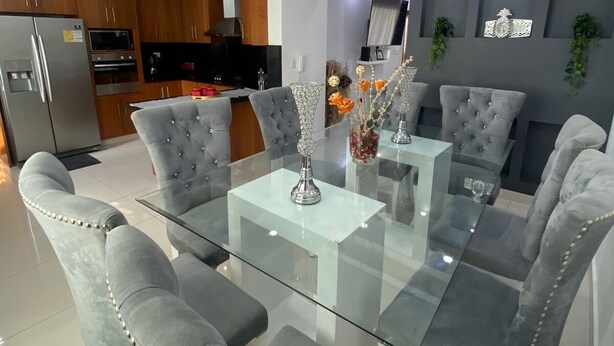 A modern dining area with a glass-top table and tufted grey chairs, a kitchen in the background, and decorative wall art. - Fully Equipped Apartment With sea View (Santo Domingo Este)