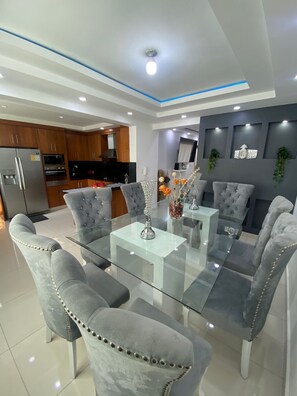 A modern dining area with a glass-top table and tufted grey chairs, a kitchen in the background, and decorative wall art.