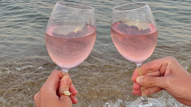Dos manos sostienen copas de vino con líquido rosado, con vista a un mar tranquilo al atardecer.