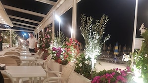 Un área de estar al aire libre con mesas y sillas blancas, rodeada de macetas con flores y cubierta por una estructura.