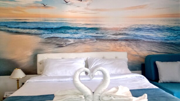 A hotel room with a bed featuring a swan towel design, a blue headboard, and a wall mural of a beach scene with birds. - Near Lake, With View, Center (Ohrid)