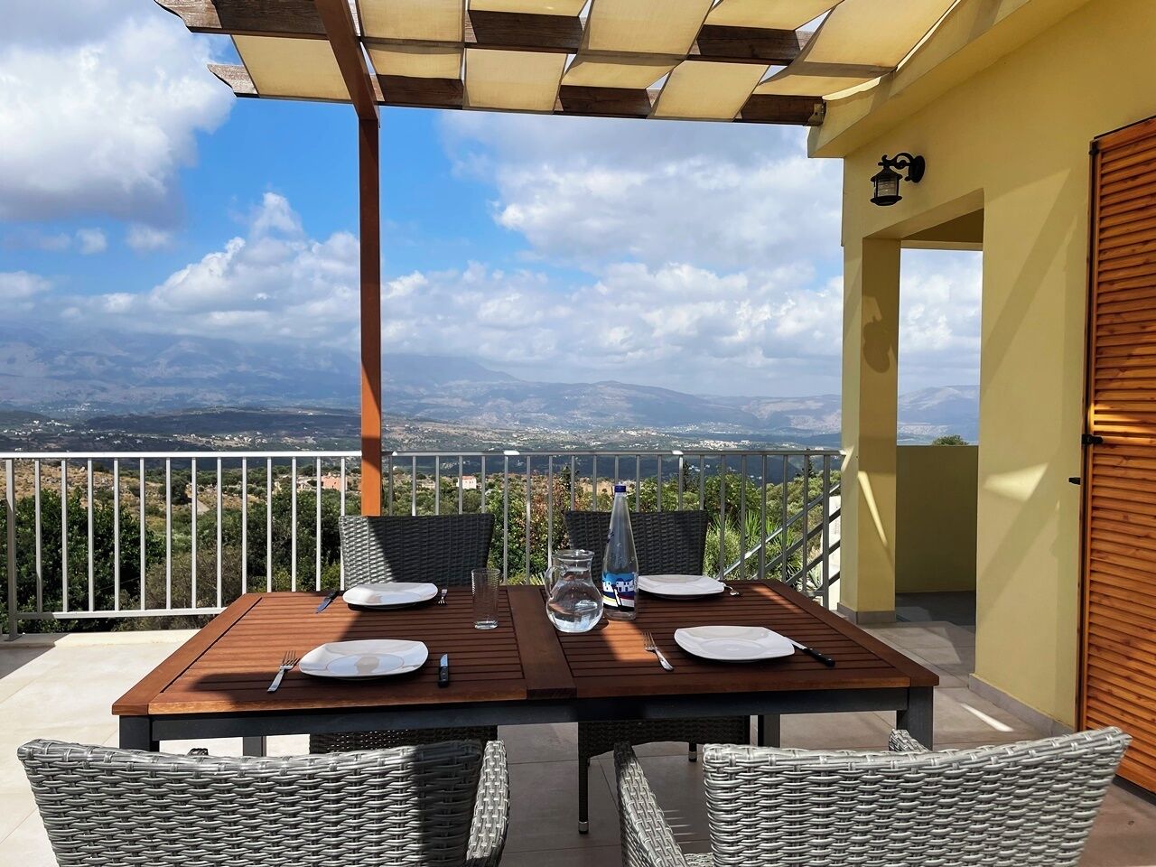 Mesa para cuatro cubierta con sillas de mimbre, una pérgola arriba y vistas a montañas distantes.