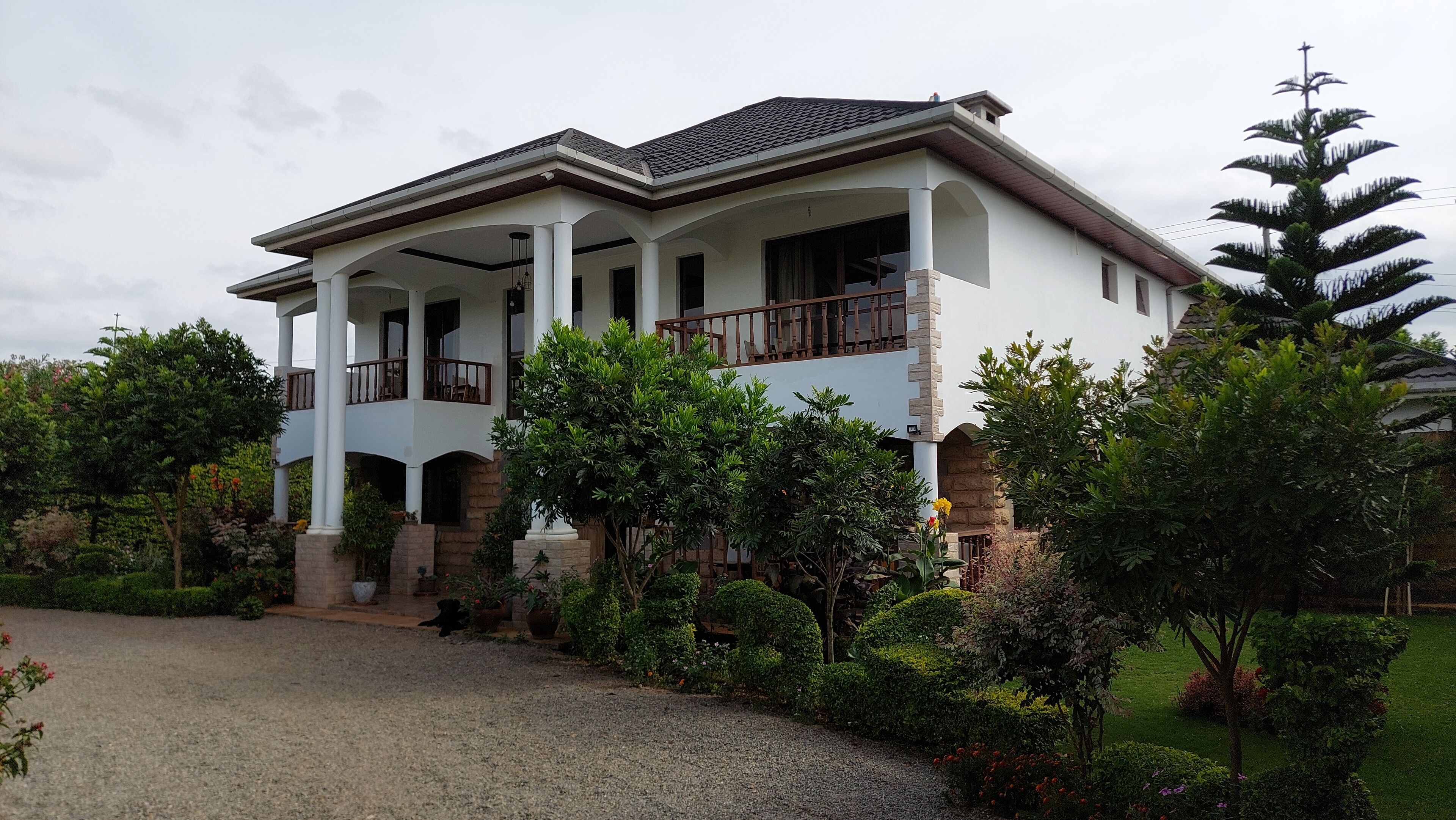 A two-story house with a balcony on the upper floor, surrounded by well-maintained gardens and a gravel driveway.
