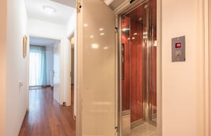 A hallway with a wooden floor, a door leading to an elevator, and a light switch on the wall.