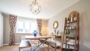 A dining area with a wooden table and bench, a bookshelf with books and decorative items, and a geometric pendant light. - Comfortable 5 Bedroom Detached House With Parking (Bicester)