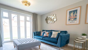 A living room with a blue sofa, a grey ottoman, a decorative wall piece, and a framed picture. - Comfortable 5 Bedroom Detached House With Parking (Bicester)