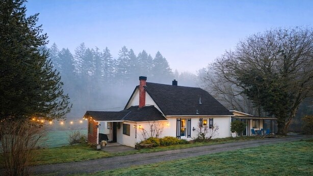 A house with a chimney, a porch with chairs, and a view of trees in the background. - 100 year old Farm Cottage. Minutes from the beach! (Nanaimo)