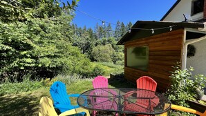 A wooden cabin with a metal table and colorful chairs in a lush garden. - 100 year old Farm Cottage. Minutes from the beach! (Nanaimo)