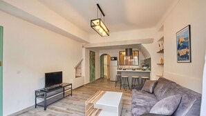 Una sala de estar moderna con un sofá gris, una mesita de centro y un televisor sobre un mueble. Se observa un área de cocina al fondo, con taburetes y una lámpara colgante.