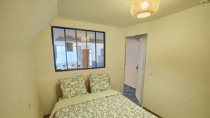 Un dormitorio con cama, ventana con vista a la cocina y puerta que da a otra habitación.