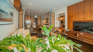 A modern living room with a sofa, coffee table, and TV stand. There is a kitchen area in the background with a dining table and chairs. - Apartment Seabreeze 1 (Canggu)