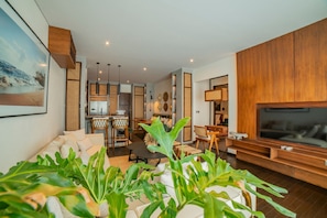 Una sala de estar moderna con sofá, mesa de centro y mueble para la televisión. Se observa un área de cocina al fondo con una mesa de comedor y sillas.