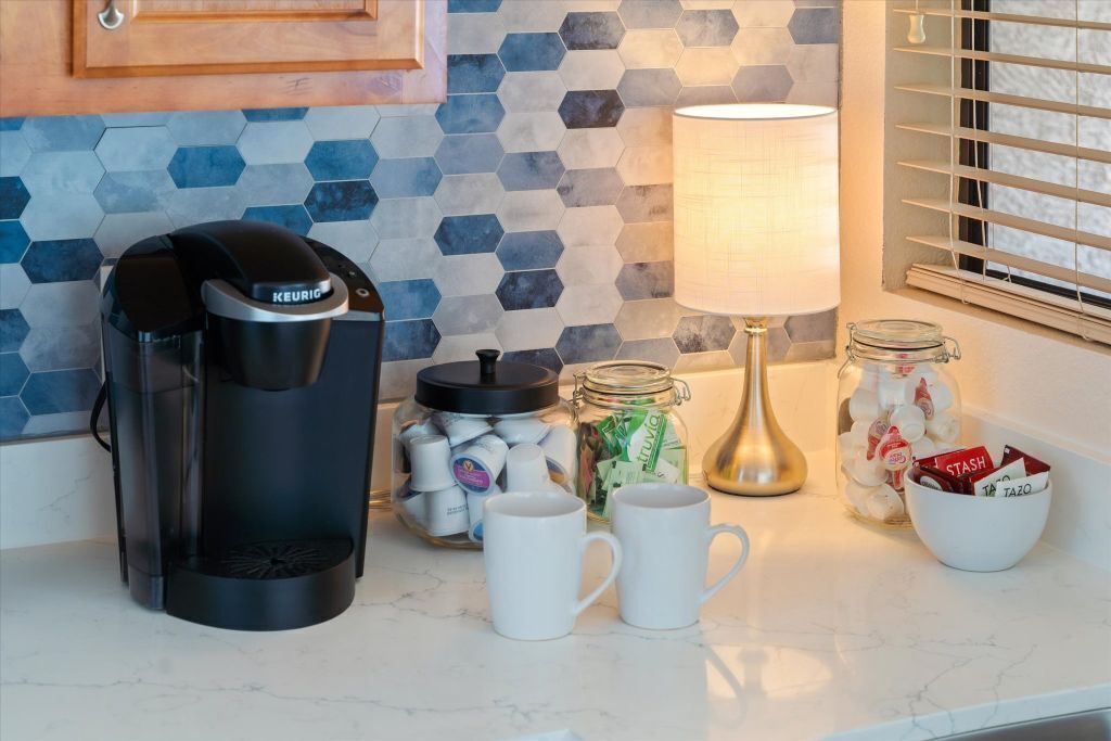 A kitchen counter with a Keurig coffee maker, jars of coffee pods, a lamp, and a bowl of snacks.