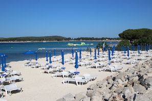 Une plage bordée de rangées de chaises longues blanches et de parasols bleus, un rivage rocheux et une mer d’un bleu limpide.