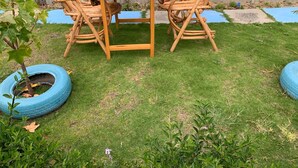 Un pequeño espacio al aire libre con una mesa de madera y dos sillas de mimbre, rodeado de un césped y una maceta de neumático azul.