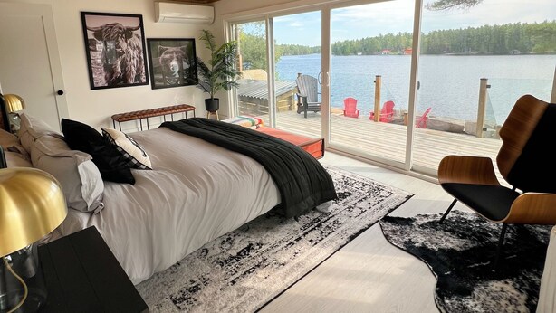 A bedroom with a large bed, a desk, a chair, and a view of a lake through large windows. - Muskoka Lakefront Resort - 5 Bedroom Modern Cottage (Muskoka Lakes)