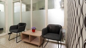 Un área de espera moderna con dos sillas negras, una mesa de madera con un florero rojo y un papel tapiz estampado.