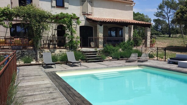 Zona junto a la piscina con una terraza de madera, sillas de descanso y un edificio de piedra con tejado de baldosas.