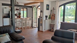 Una sala de estar con un sofá de cuero negro, un sillón gris y un comedor con sillas blancas. Un ventanal ofrece vistas al exterior.