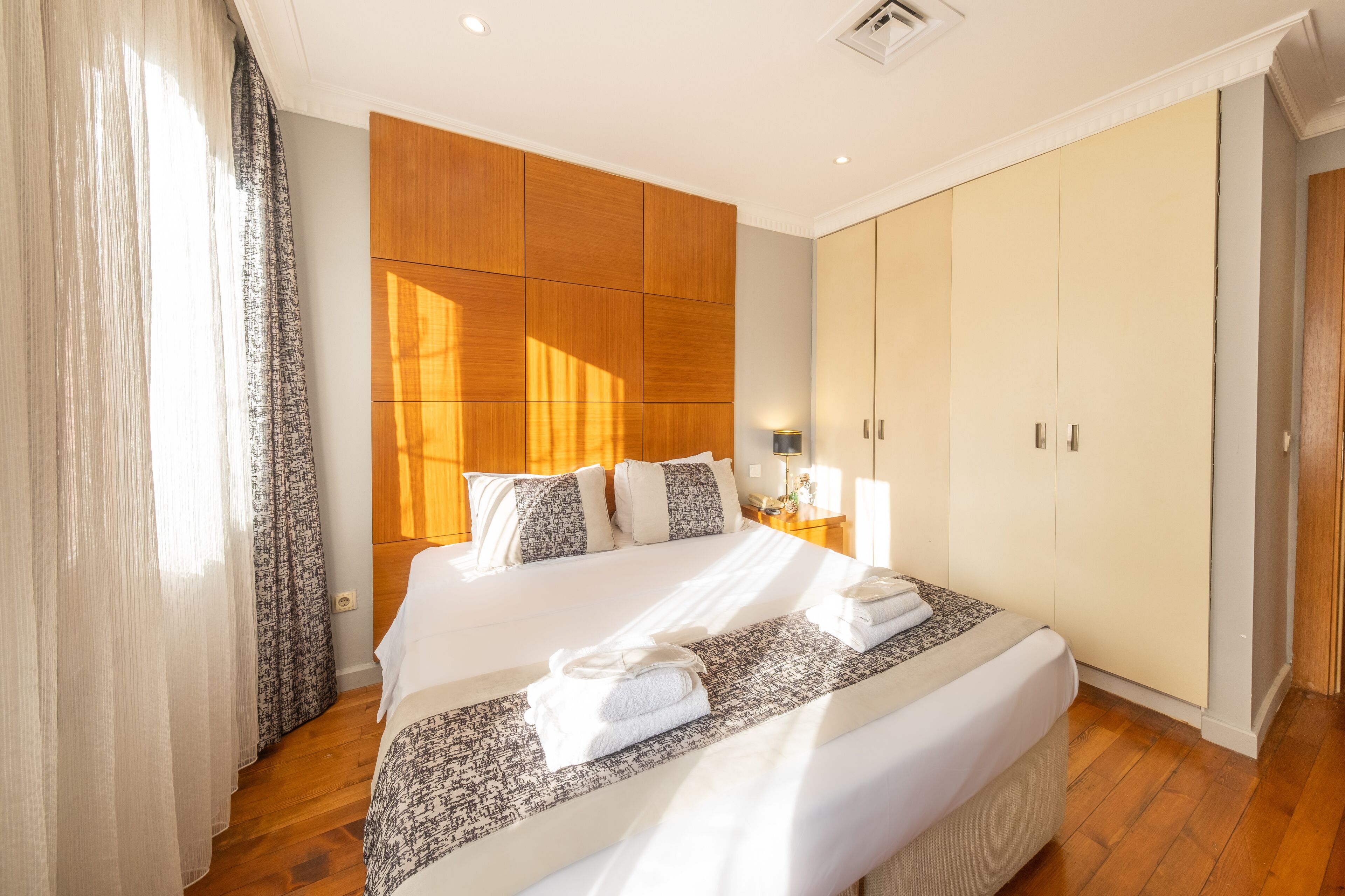 A hotel room with a large bed, wooden wardrobe, and a bedside table with a lamp.