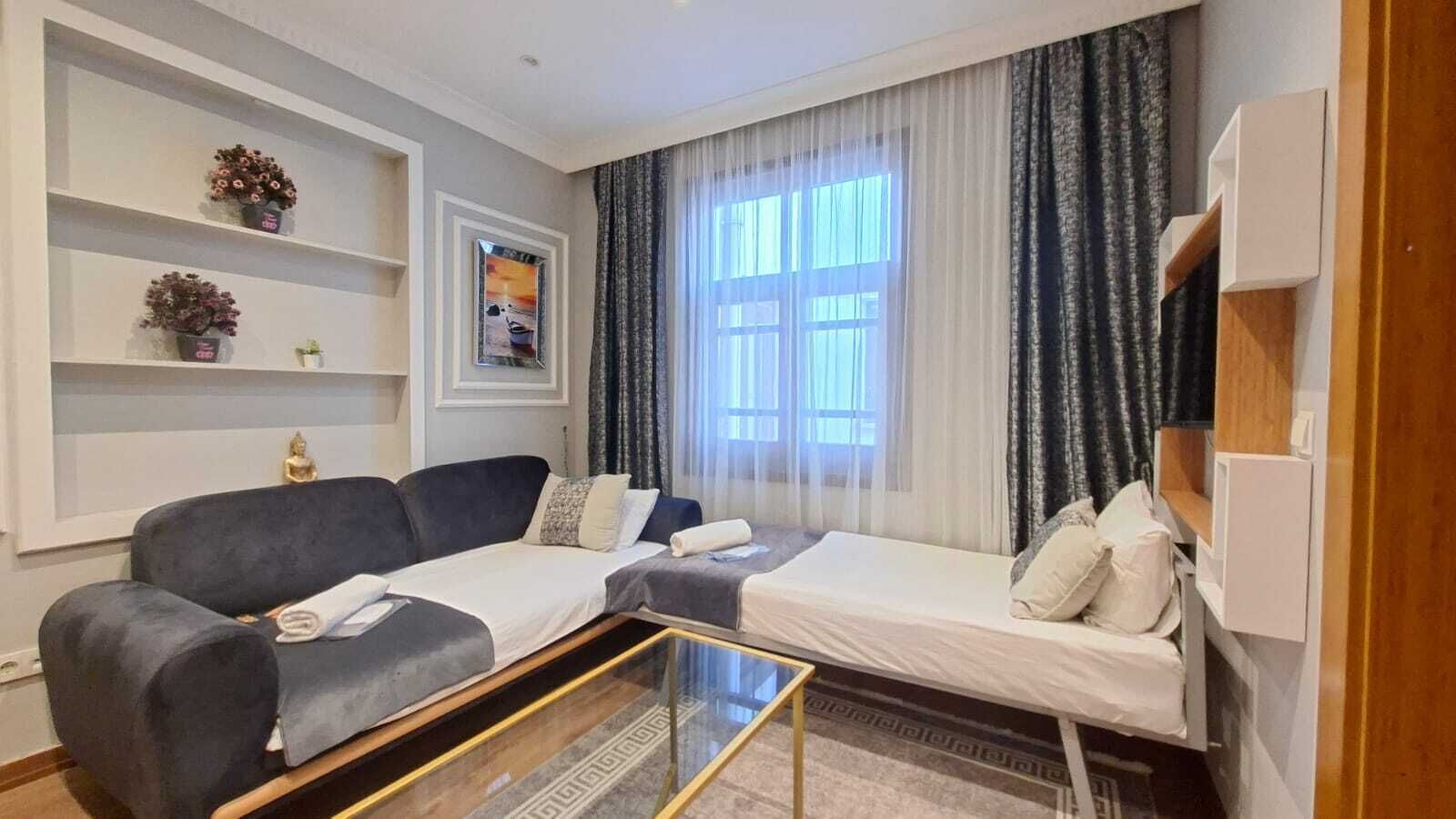Una sala de estar moderna con un sofá, una cama más pequeña, una mesa de centro de vidrio y un ventanal con cortinas.