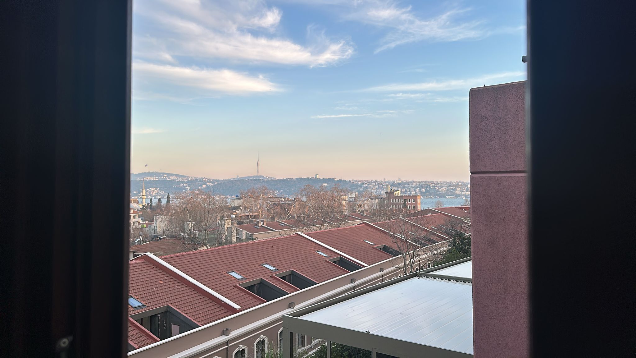 A view from a window overlooking a cityscape with red-roofed buildings and a distant tower.