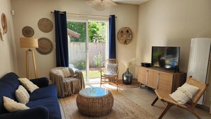 Ein Wohnzimmer mit Sofa, Sessel, Fernseher und einem großen Fenster mit Vorhängen.