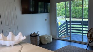 A modern living room with a large white rug, a white decorative bowl, a woven basket, and a beige ottoman. A flat-screen TV is mounted on the wall, and there is a sliding glass door leading to an outdoor deck.