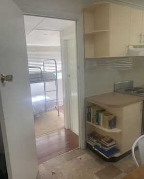 A compact kitchen with a built-in stove, a small bookshelf with books, and a bunk bed in the adjacent room. - Comfortable Family Snow Accommodation. (Talbingo)