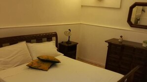 Una cama bien hecha con sábanas blancas y dos almohadones decorativos, una mesita de noche con lámpara, un aparador y un espejo montado en la pared.