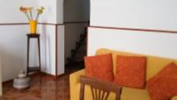 Una sala de estar con un sofá amarillo, una pequeña mesa de madera y un florero con flores sobre una mesita auxiliar.