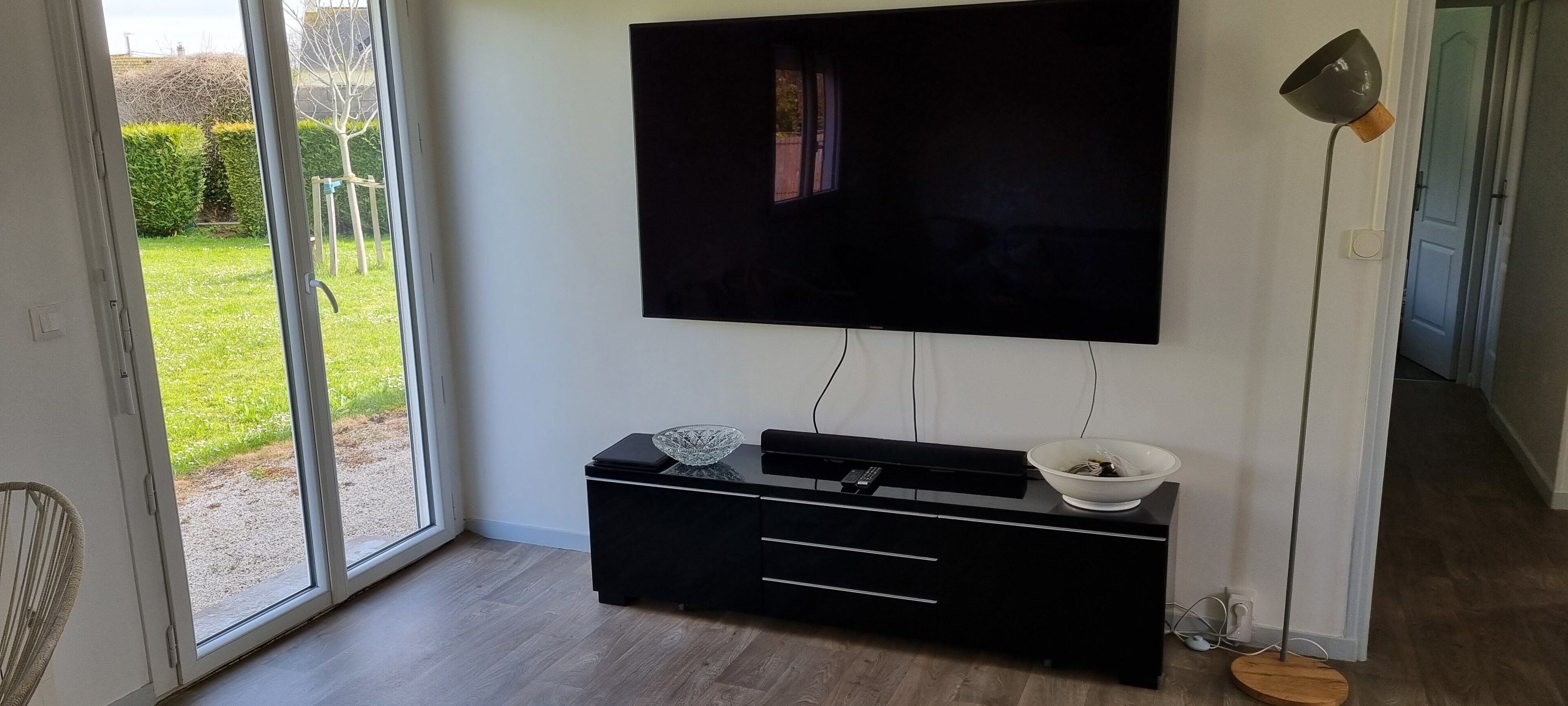 A modern living room with a flat-screen TV mounted on the wall, a black entertainment unit with drawers, a glass bowl, and a floor lamp.