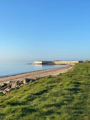Un paysage côtier comprenant une zone herbeuse, une plage de sable et un mur de pierres qui s’étend dans l’eau.