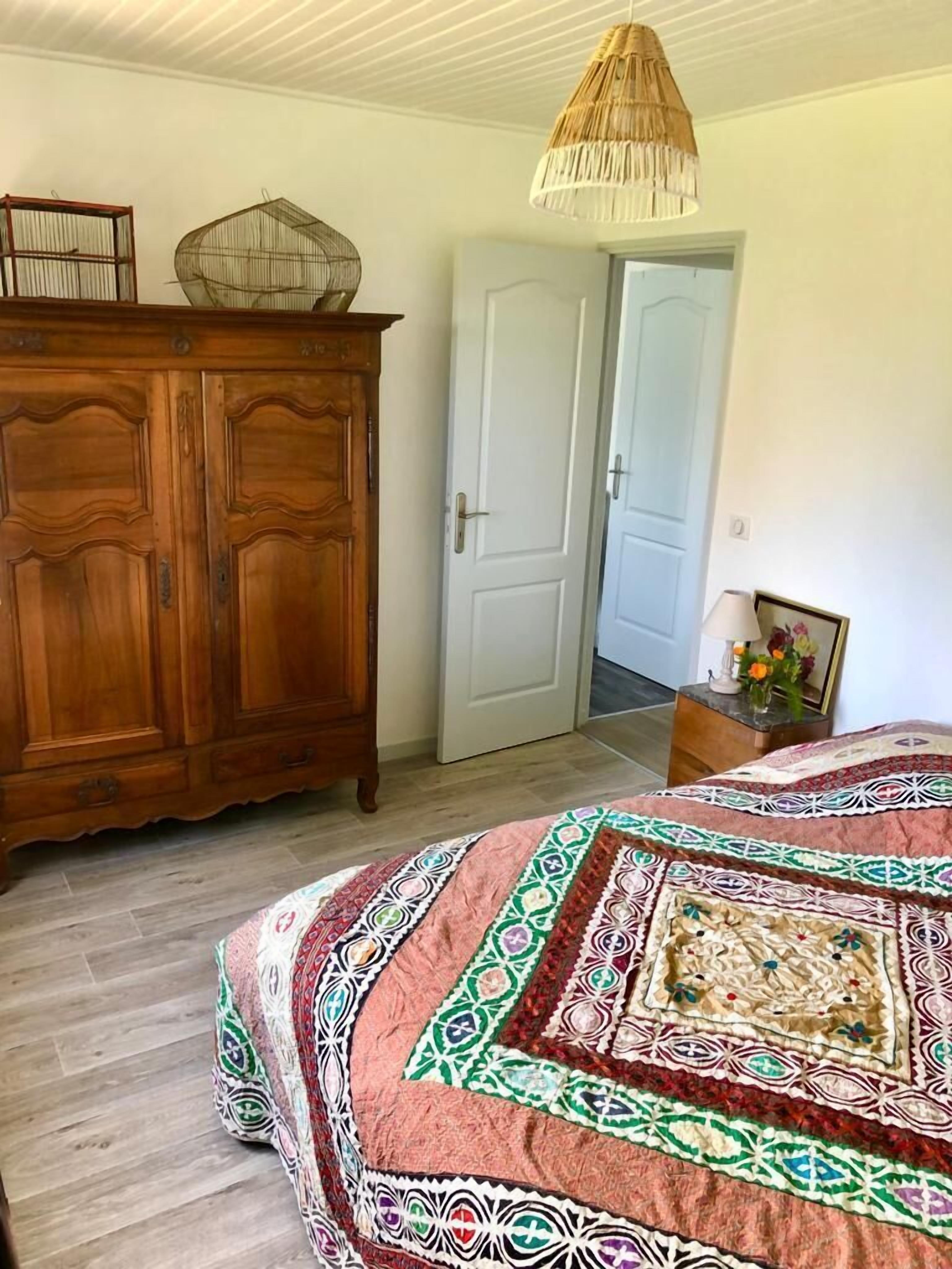 A bedroom with a wooden wardrobe, a bed with a patterned bedspread, a hanging light fixture, and a door leading to another room.