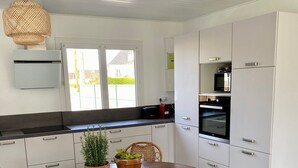 A modern kitchen with white cabinets, a wooden dining table, wicker chairs, and a hanging light fixture. - The Saint Vaast orchard  (Saint-Vaast-la-Hougue)