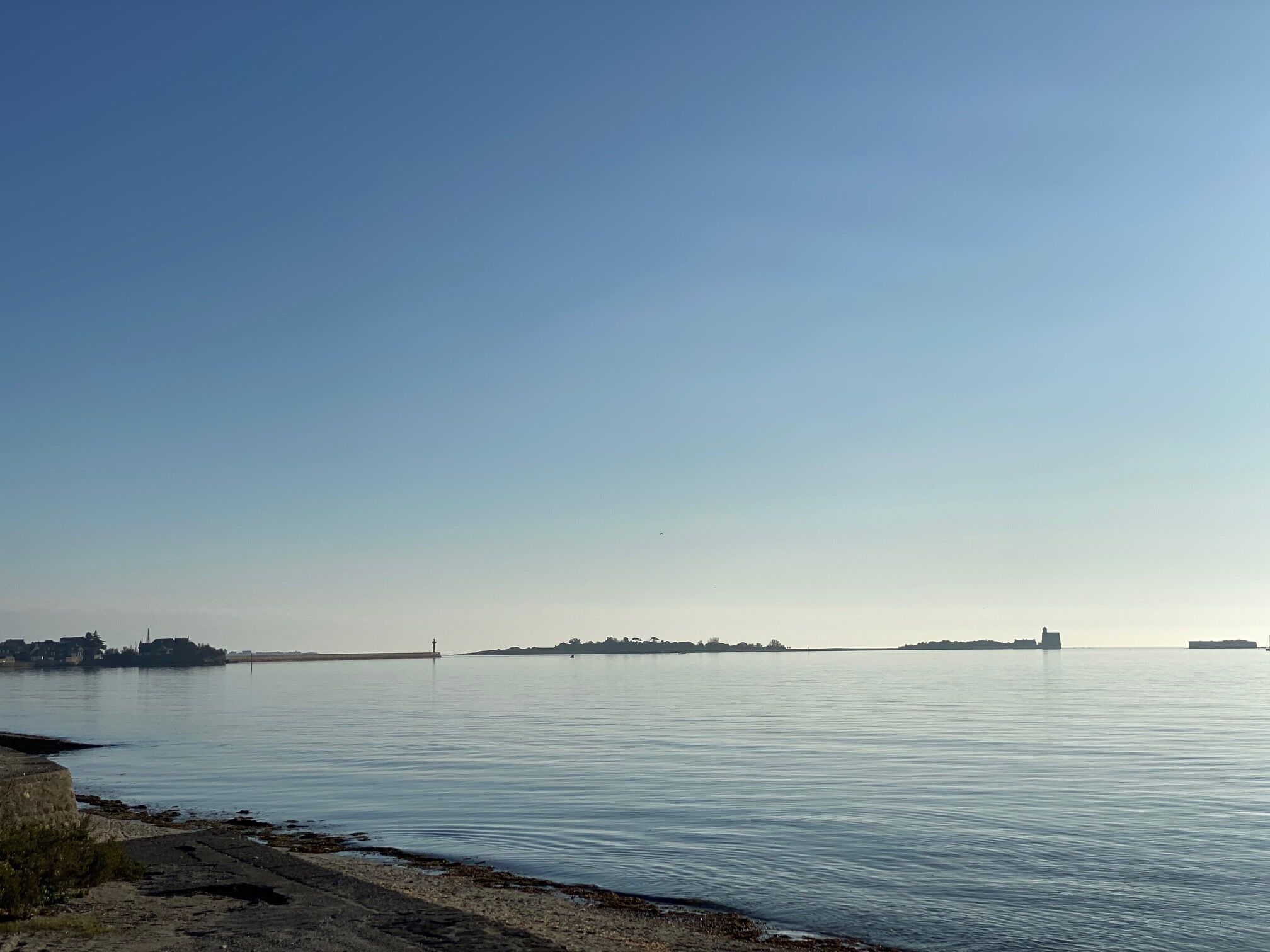 Un front marítim tranquil amb cel serè, una platja sorrenca i edificis al lluny.